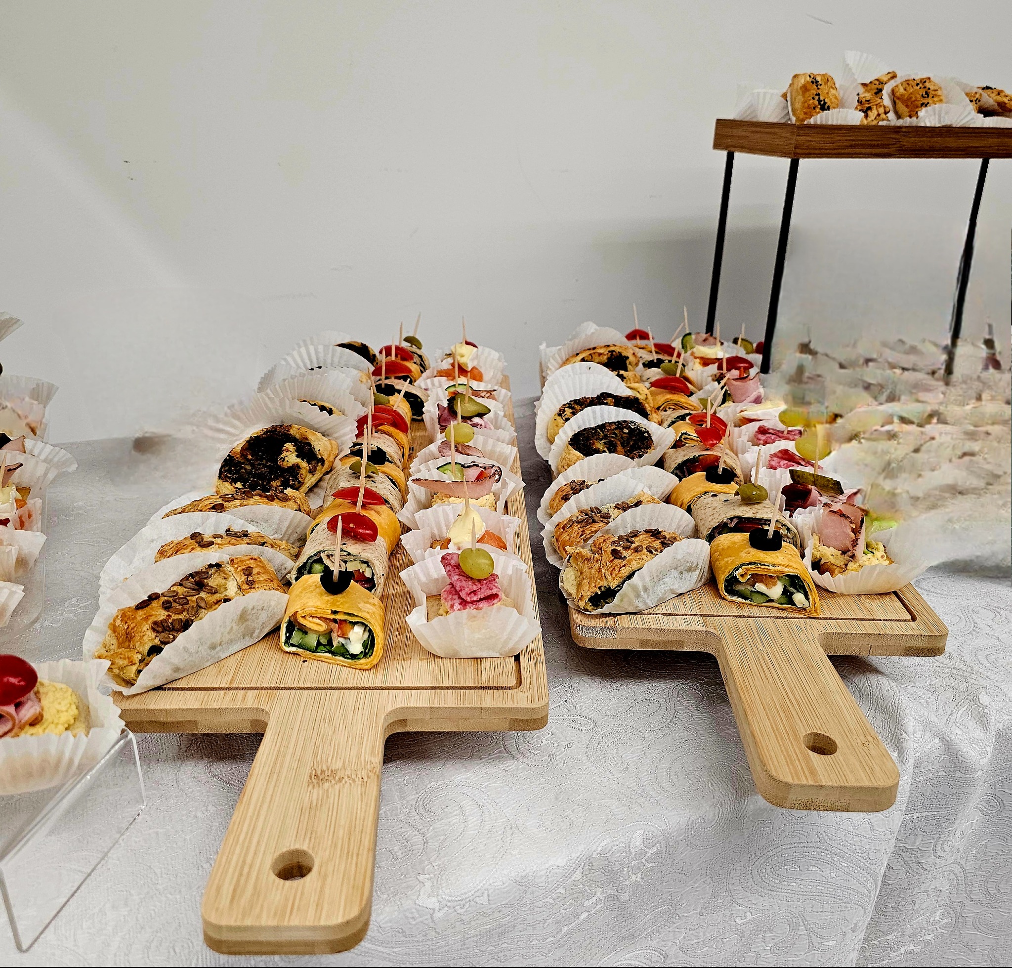 Ekskluzywny catering: deski z roladkami, tartaletkami i przekąskami na stole z białym obrusem, w tle bufet z ciastkami. Elegancka prezentacja.