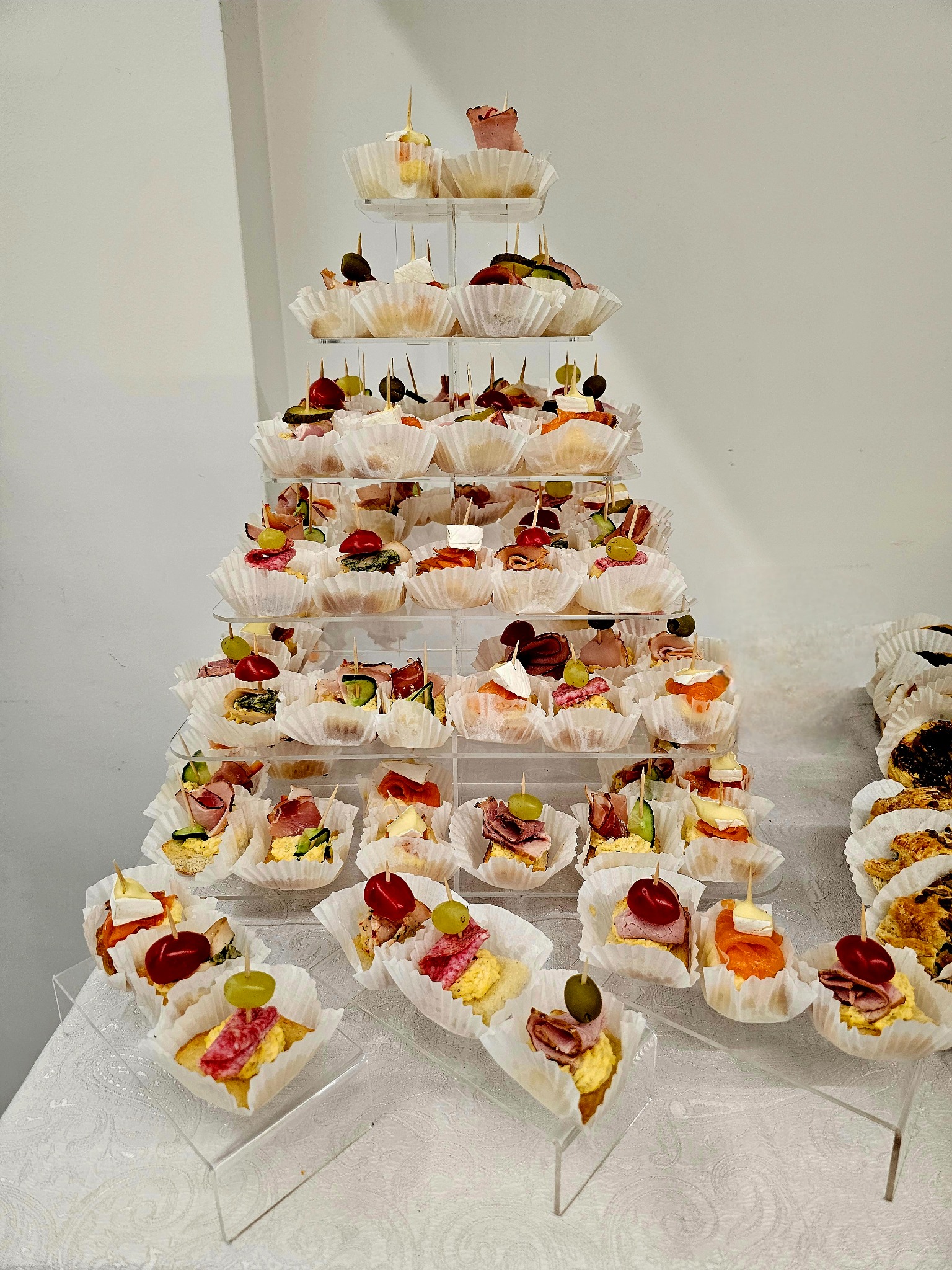 Piramida z apetycznych kanapek na przezroczystych półkach, każda w papierowej foremce, ozdobiona oliwkami, winogronami i wędlinami. Elegancka prezentacja przekąsek.