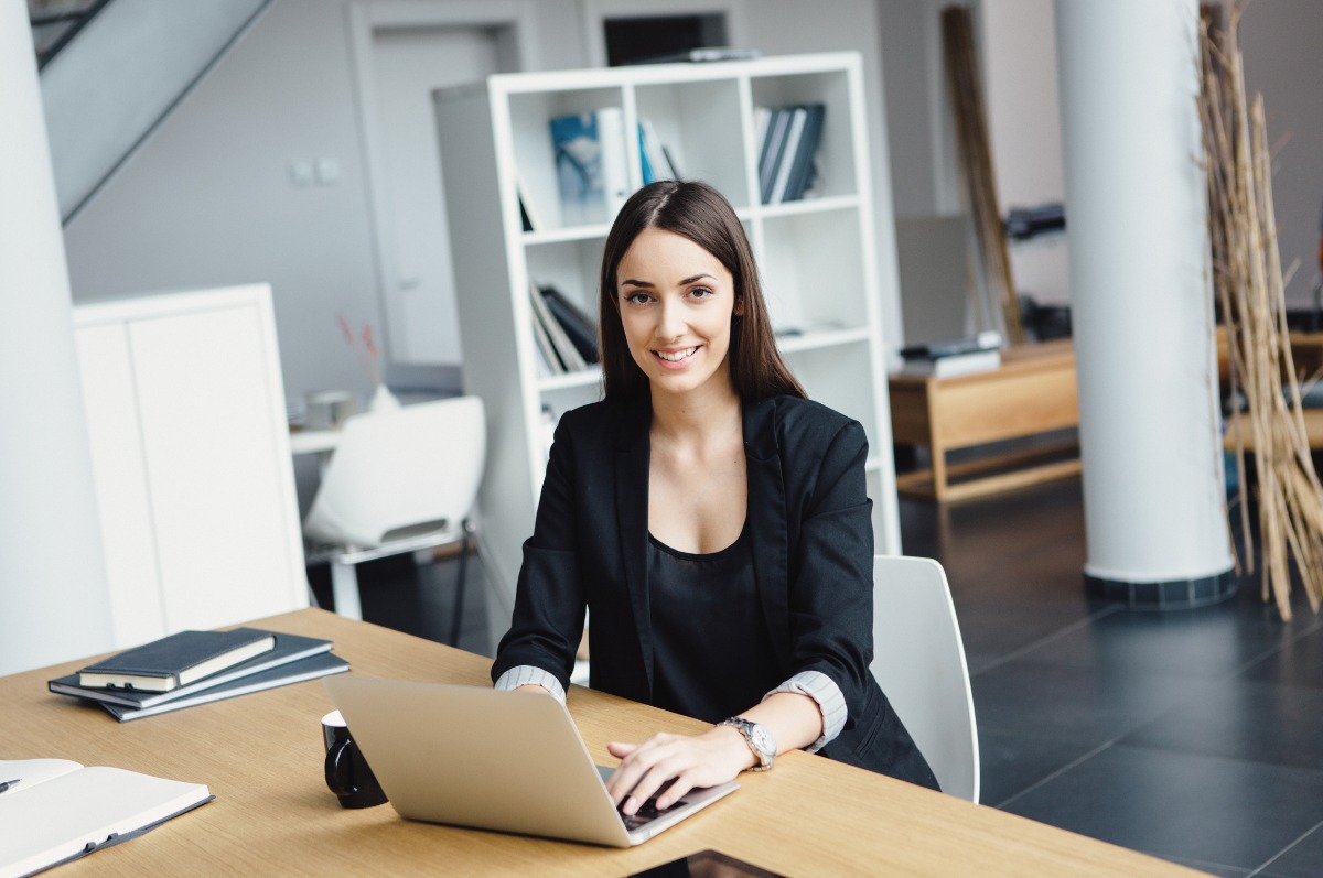 Uśmiechnięta kobieta w biurze, w czarnej marynarce, pracuje na laptopie. Na biurku notatniki i kawa. W tle regał z książkami i kolumna.