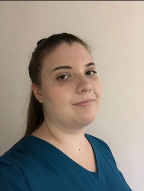 Portret kobiety w turkusowym uniformie, z włosami upiętymi w kok i długim kucykiem, uśmiechniętej, na tle jasnej ściany, z delikatnym makijażem.