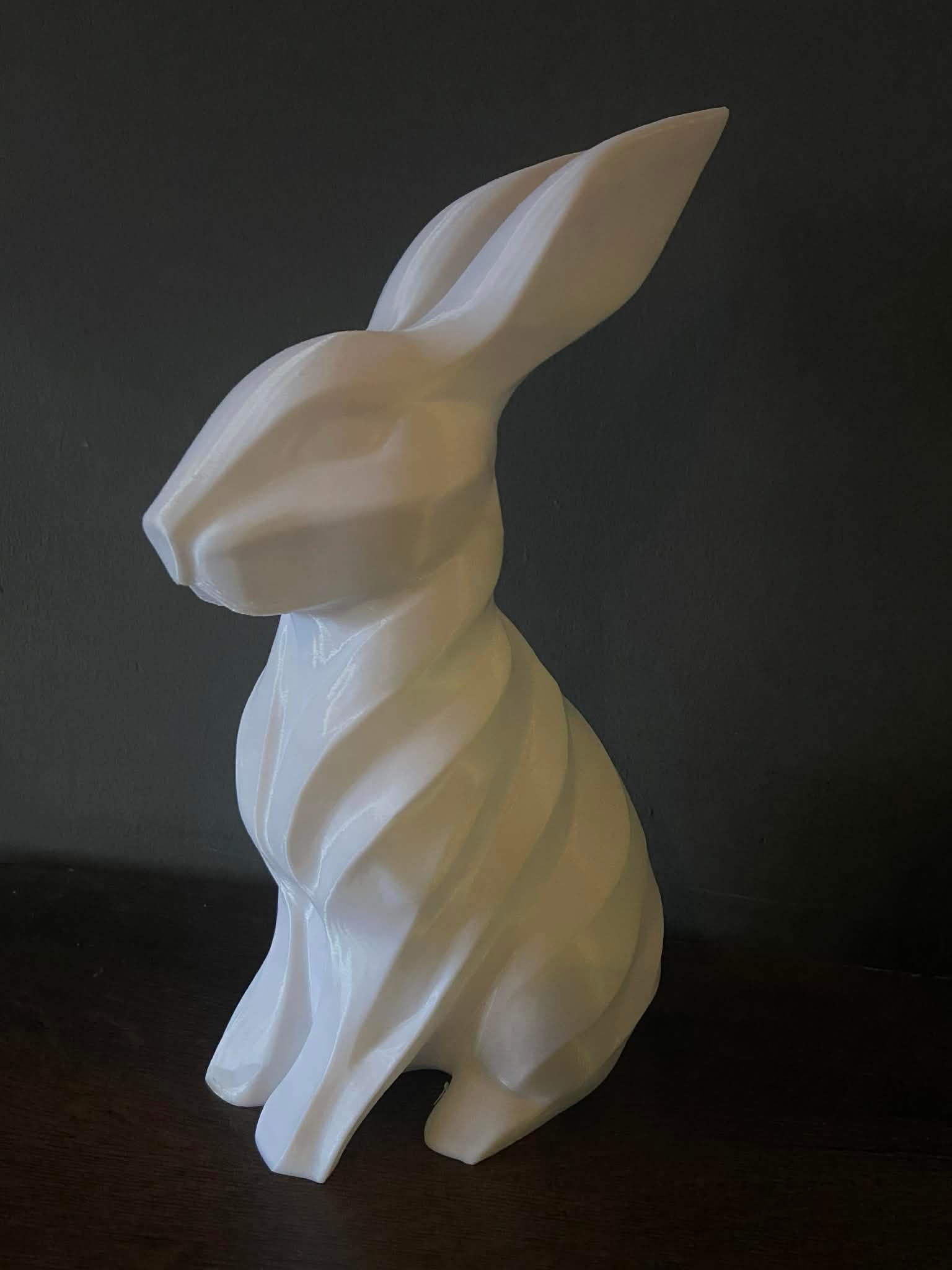 Biała, spiralna figurka królika, wydrukowana w 3D, na ciemnym tle. Widoczne warstwy druku, nadające teksturę. Stoi na ciemnym blacie.