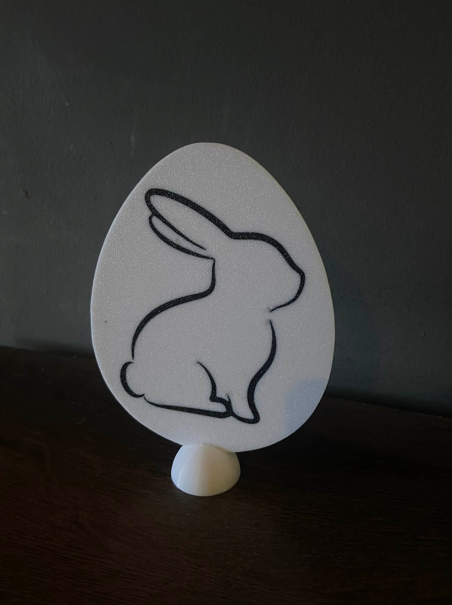 Biała, jajowata dekoracja z czarnym konturem królika, wydrukowana w 3D w Ustroniu. Stoi na małej, okrągłej podstawie na drewnianej powierzchni.