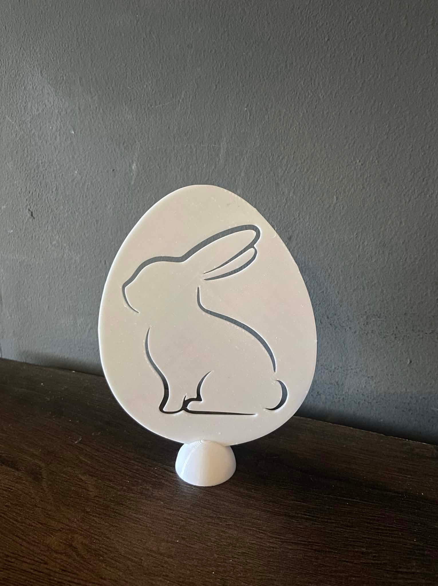 Biała, trójwymiarowa ozdoba w kształcie jajka z wyciętym zarysem królika, umieszczona na małej, okrągłej podstawce. Dekoracja stoi na ciemnym, drewnianym blacie.