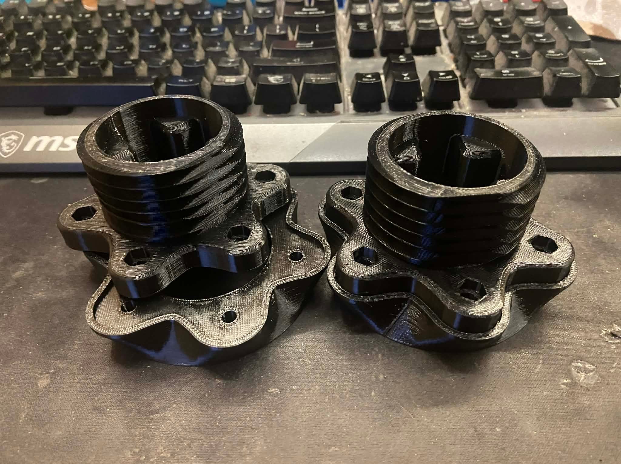 Dwa czarne, identyczne elementy z tworzywa sztucznego, wydrukowane w technologii 3D, leżące na biurku z klawiaturą w tle. Widoczne warstwy druku.