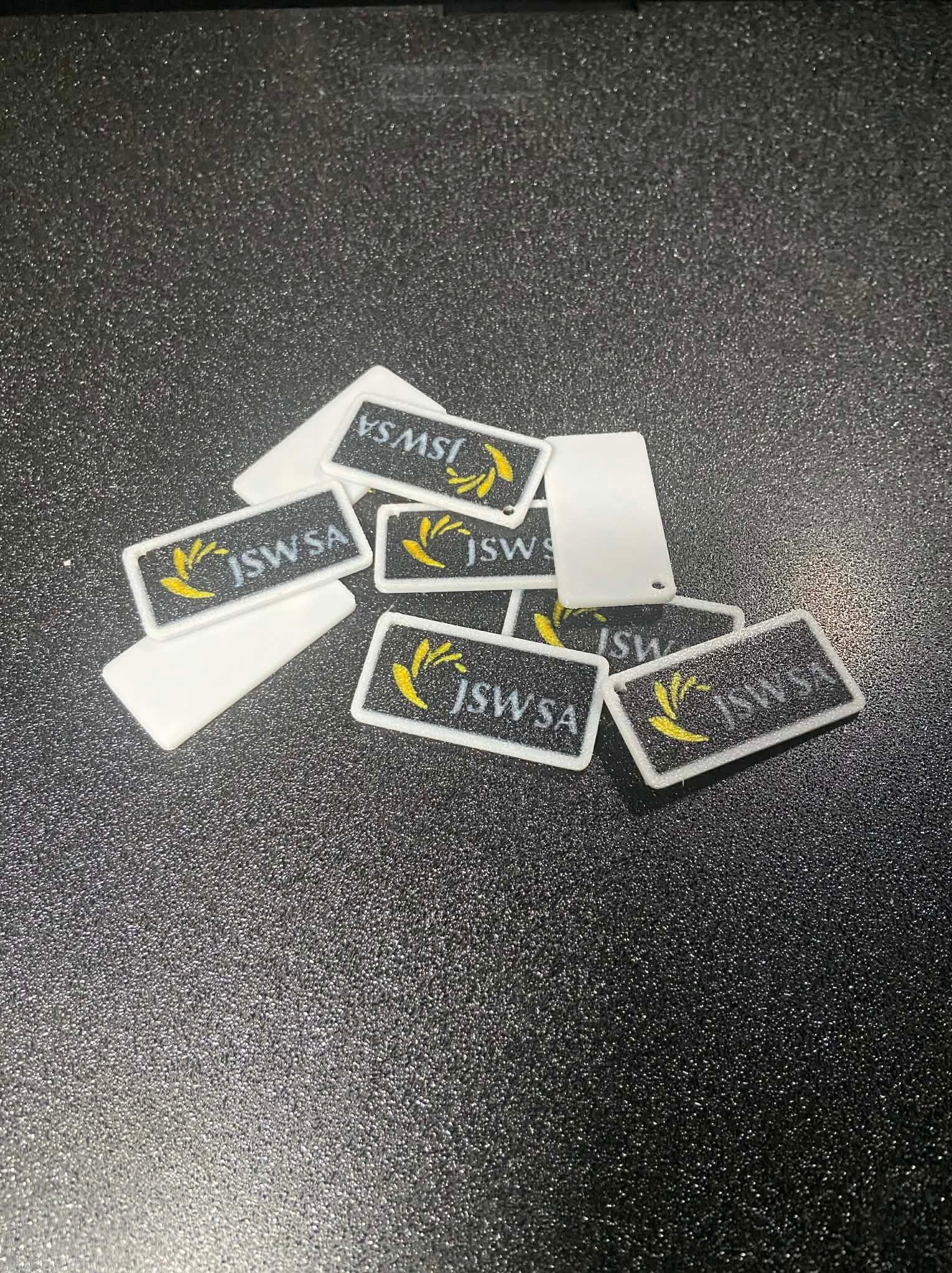 Partia białych, prostokątnych breloków z logo JSW SA na czarnym, błyszczącym tle. Breloki wykonane z tworzywa sztucznego, z widoczną fakturą.