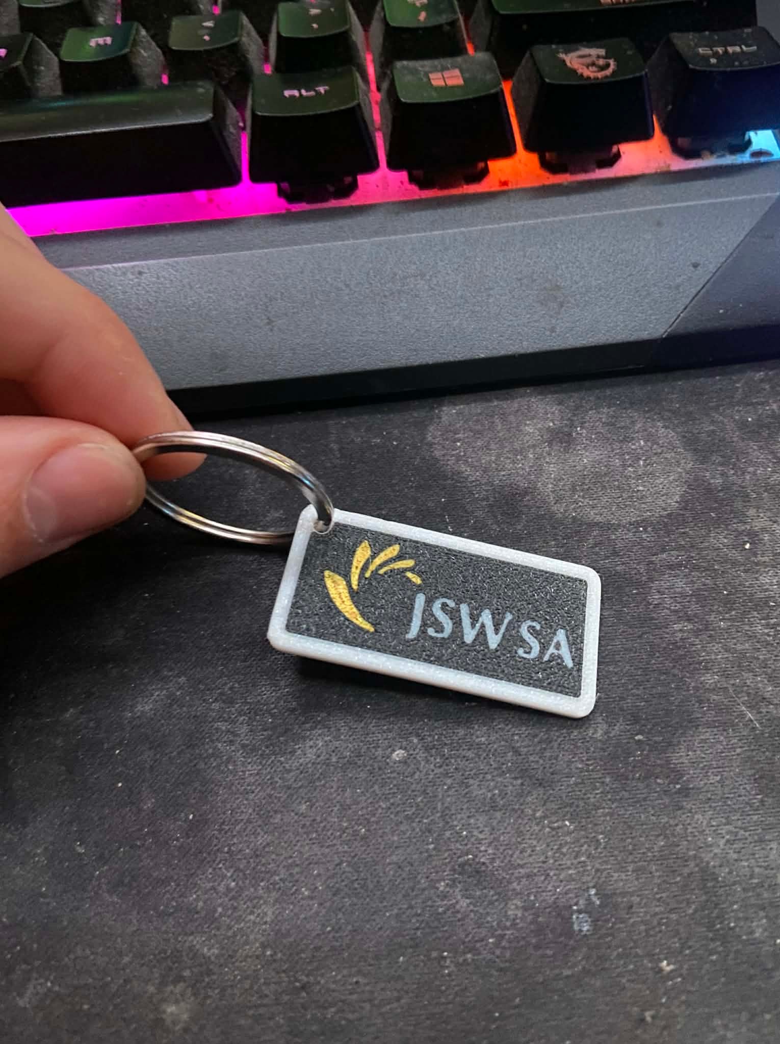 Brelok z logo JSW SA, wydrukowany w 3D, trzymany w dłoni na tle klawiatury gamingowej z podświetleniem RGB. Detaliczne ujęcie prezentuje jakość druku i precyzję wykonania.