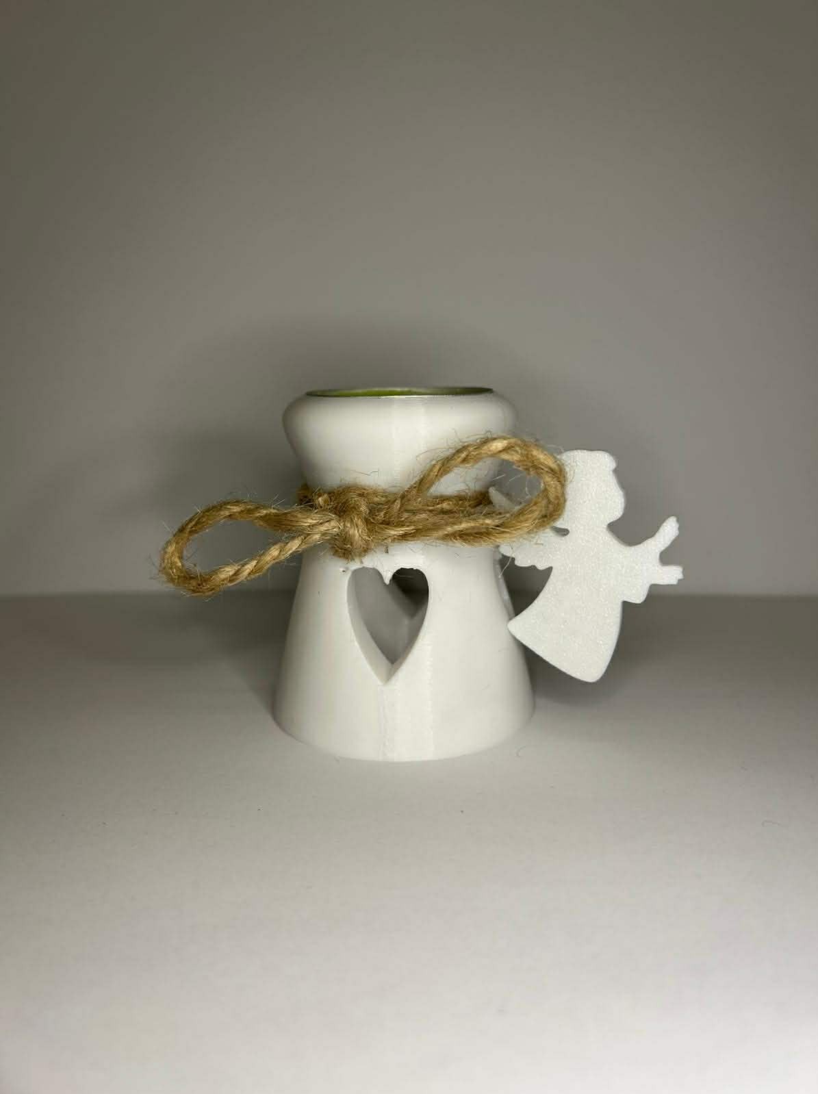 Biały, stożkowy świecznik z sercem, ozdobiony jutowym sznurkiem i aniołkiem, wykonany prawdopodobnie metodą druku 3D. Widoczne warstwy druku.