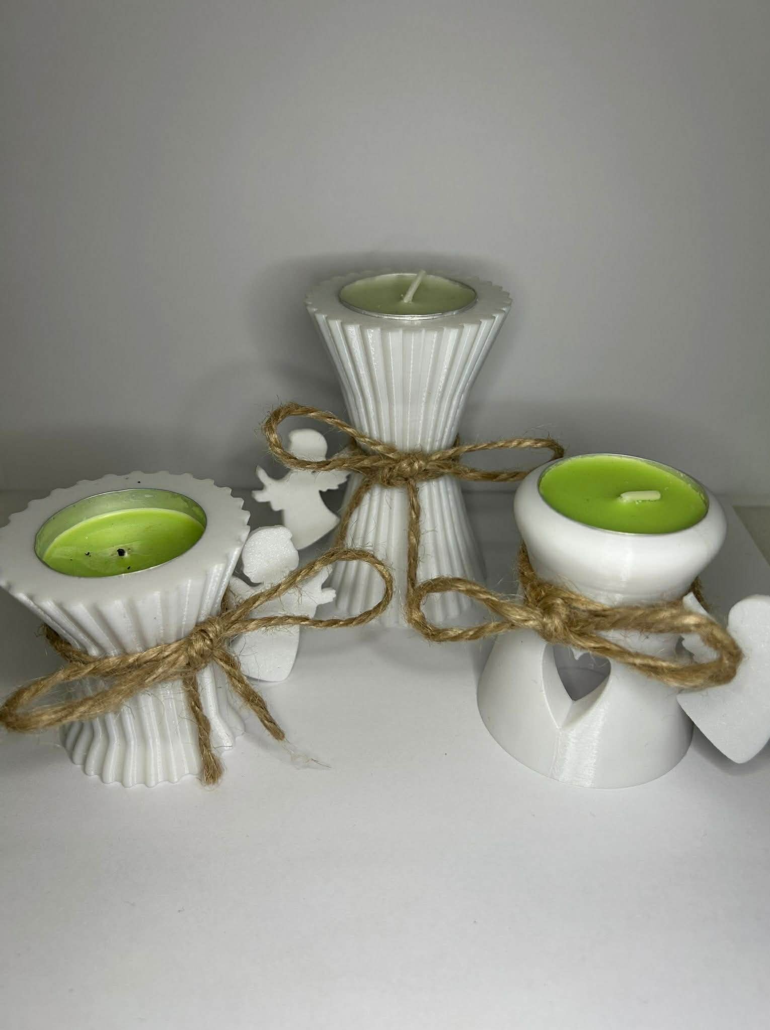 Trzy białe świeczniki z zielonymi świecami, wykonane metodą druku 3D, ozdobione sznurkiem jutowym i białymi figurkami aniołków, na jasnym tle.