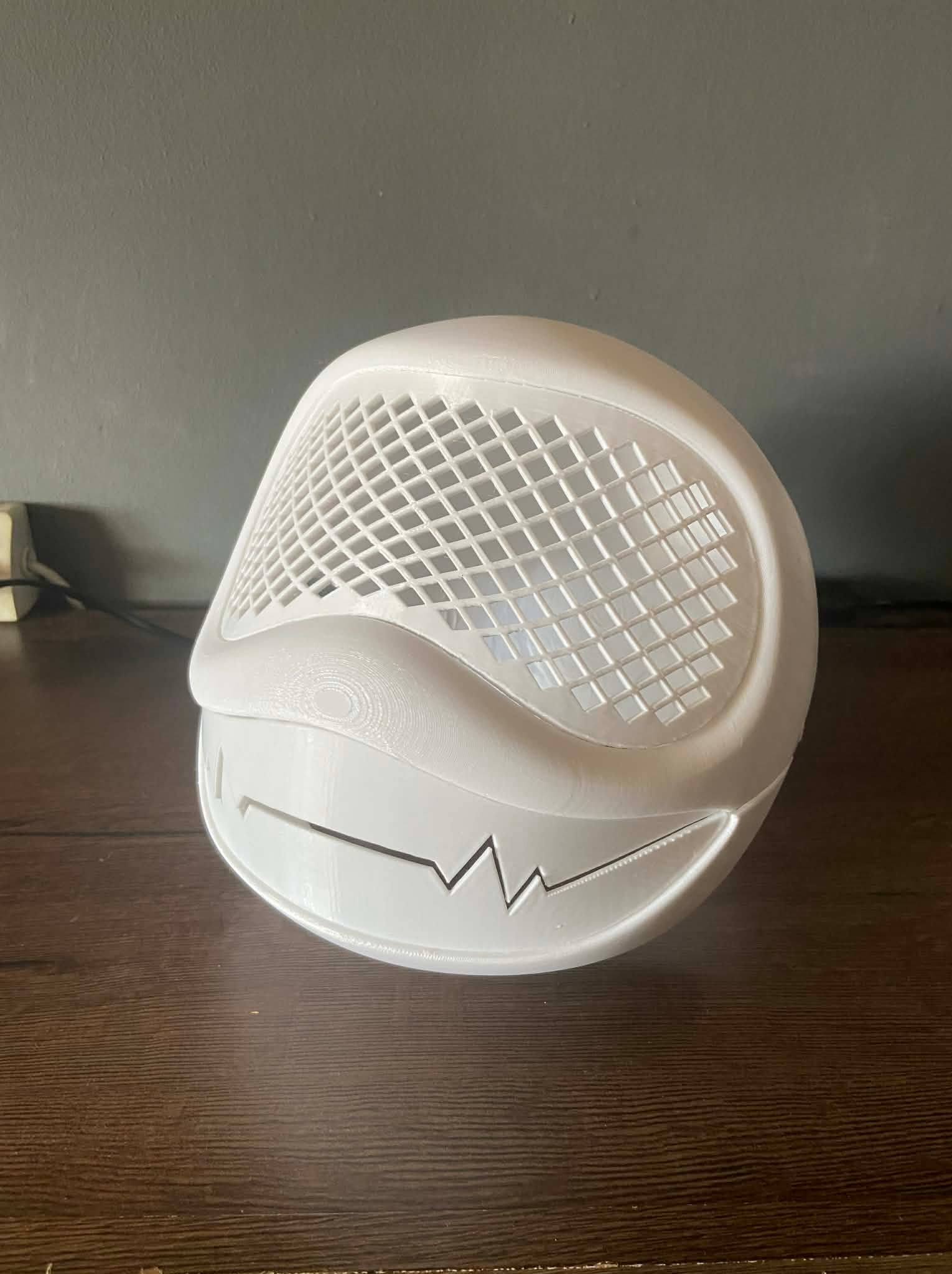 Biały, trójwymiarowy model maski z siatką i wyciętym wzorem uśmiechu, wydrukowany na drukarce 3D, leżący na drewnianym blacie na tle szarej ściany.