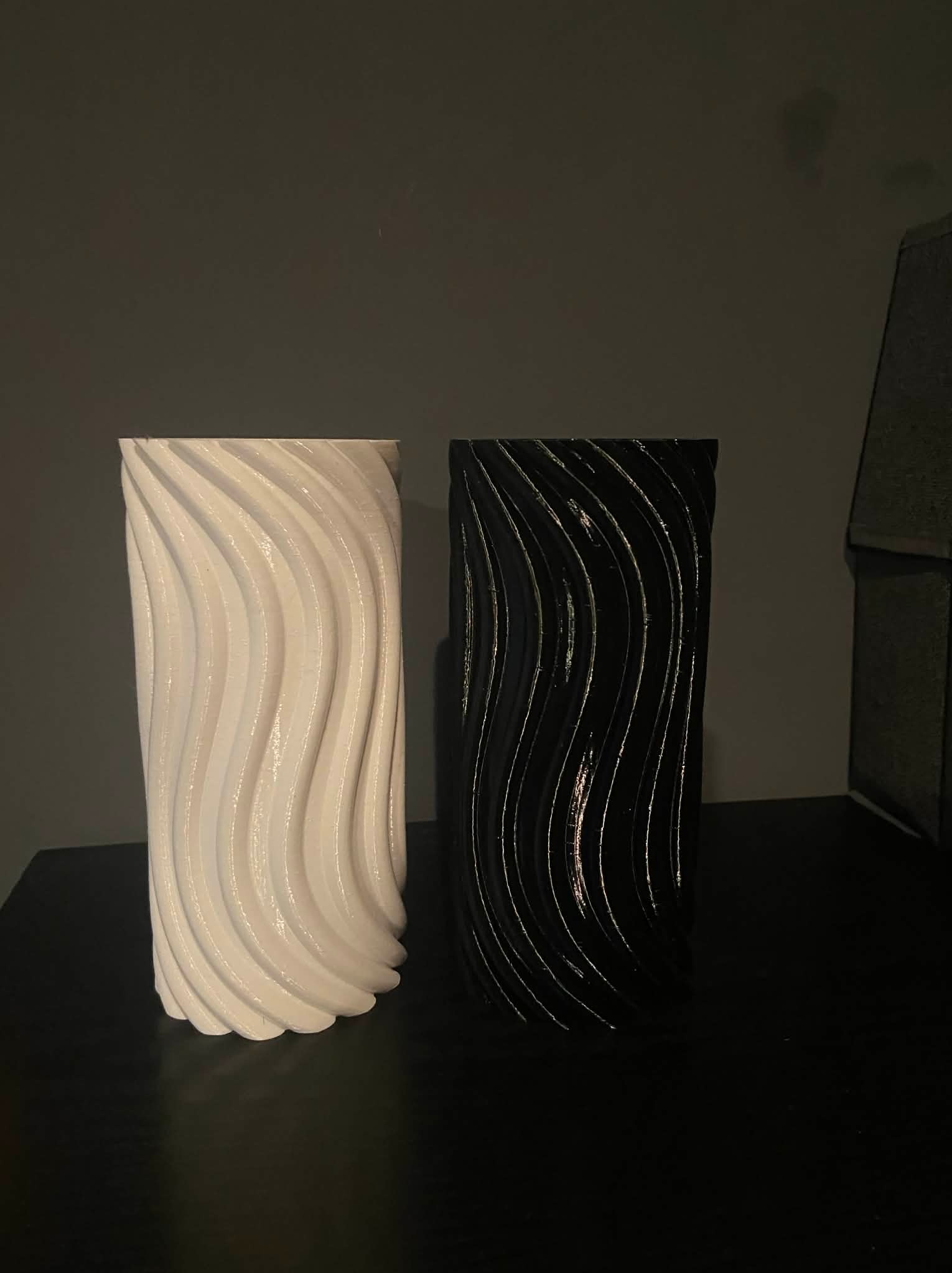 Dwa wazony z wydruku 3D: biały i czarny, z falistą teksturą, stoją obok siebie na ciemnej powierzchni. Minimalistyczny design i kontrast kolorów.
