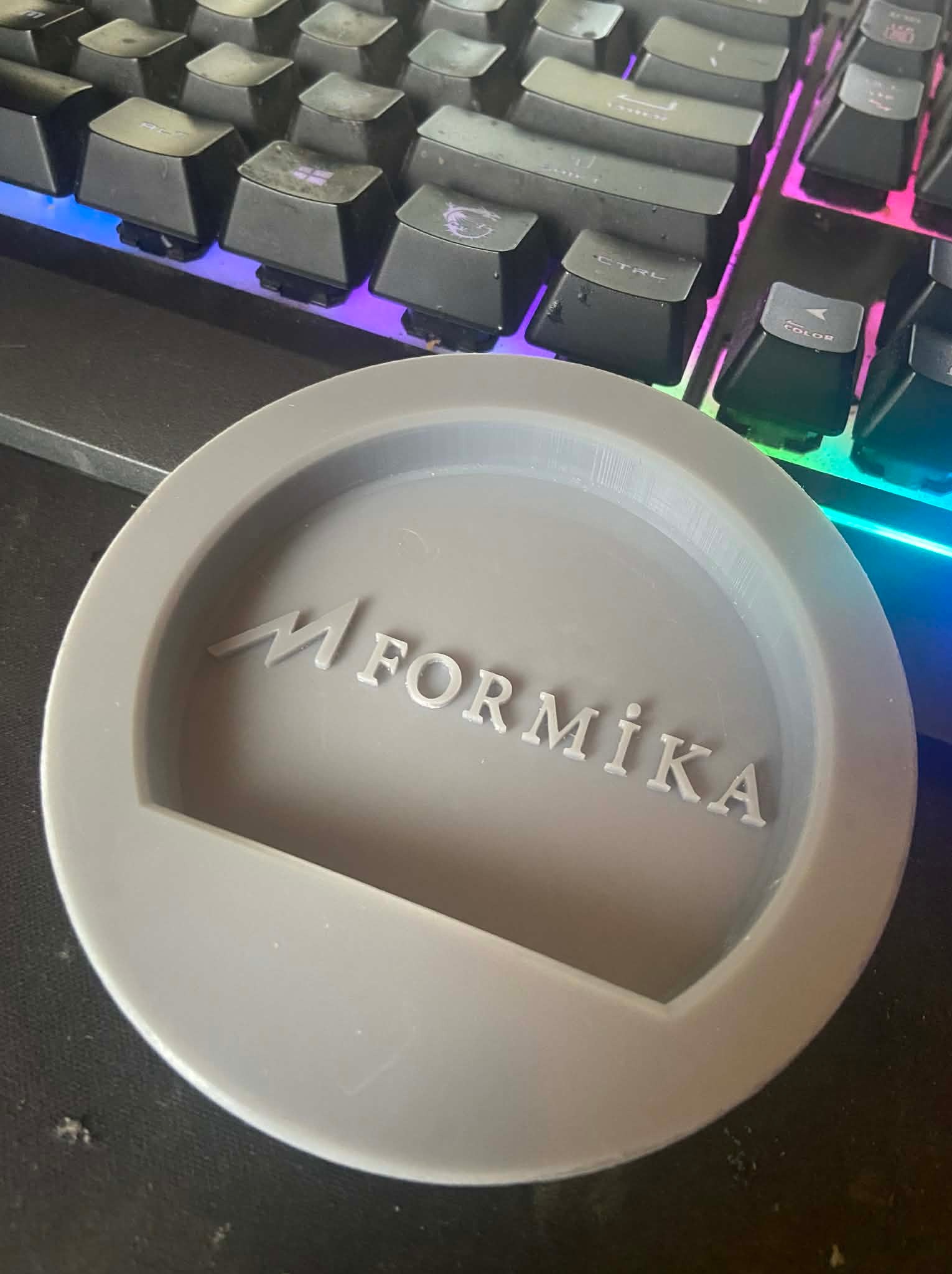 Szary, okrągły podstawek z napisem 'M Formika' leży na biurku obok podświetlanej klawiatury. Widoczne ślady użytkowania na biurku.