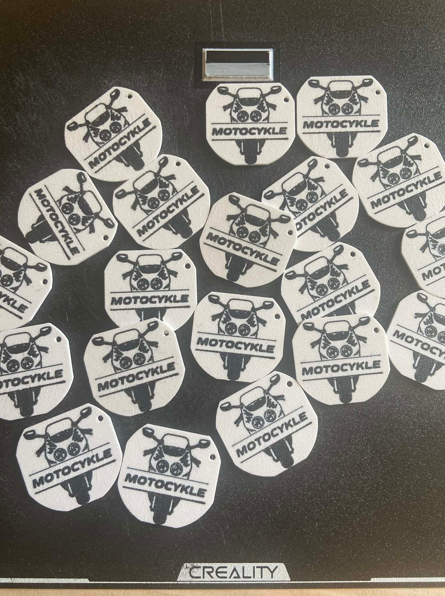Partia białych, okrągłych zawieszek z motywem motocykla i napisem 'MOTOCYKLE', wykonanych prawdopodobnie metodą druku 3D, leżących na czarnej powierzchni.