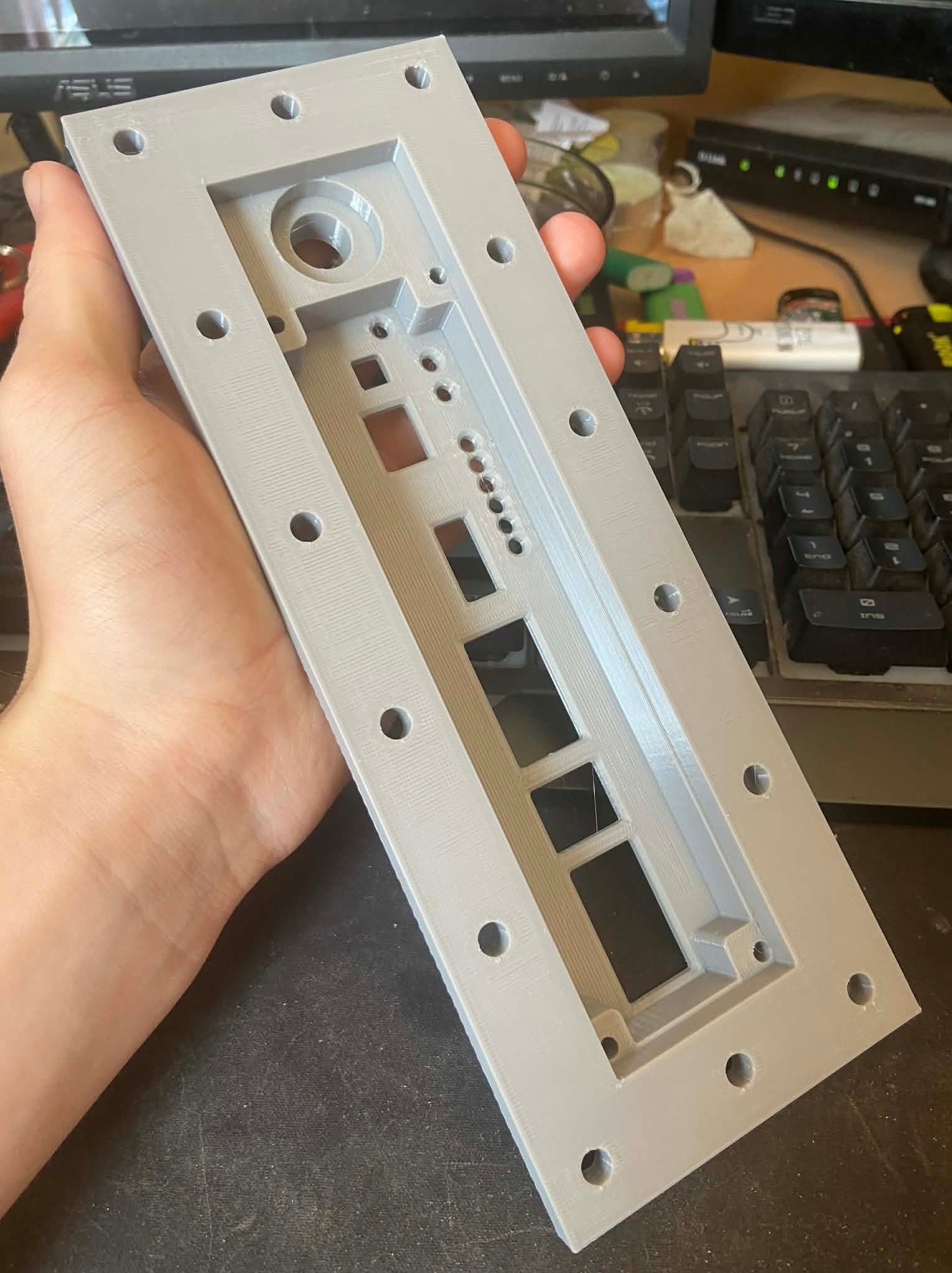 Szary, wydrukowany w 3D element obudowy z otworami i wgłębieniami, trzymany w dłoni na tle biurka z monitorem i klawiaturą. Widoczna tekstura druku.