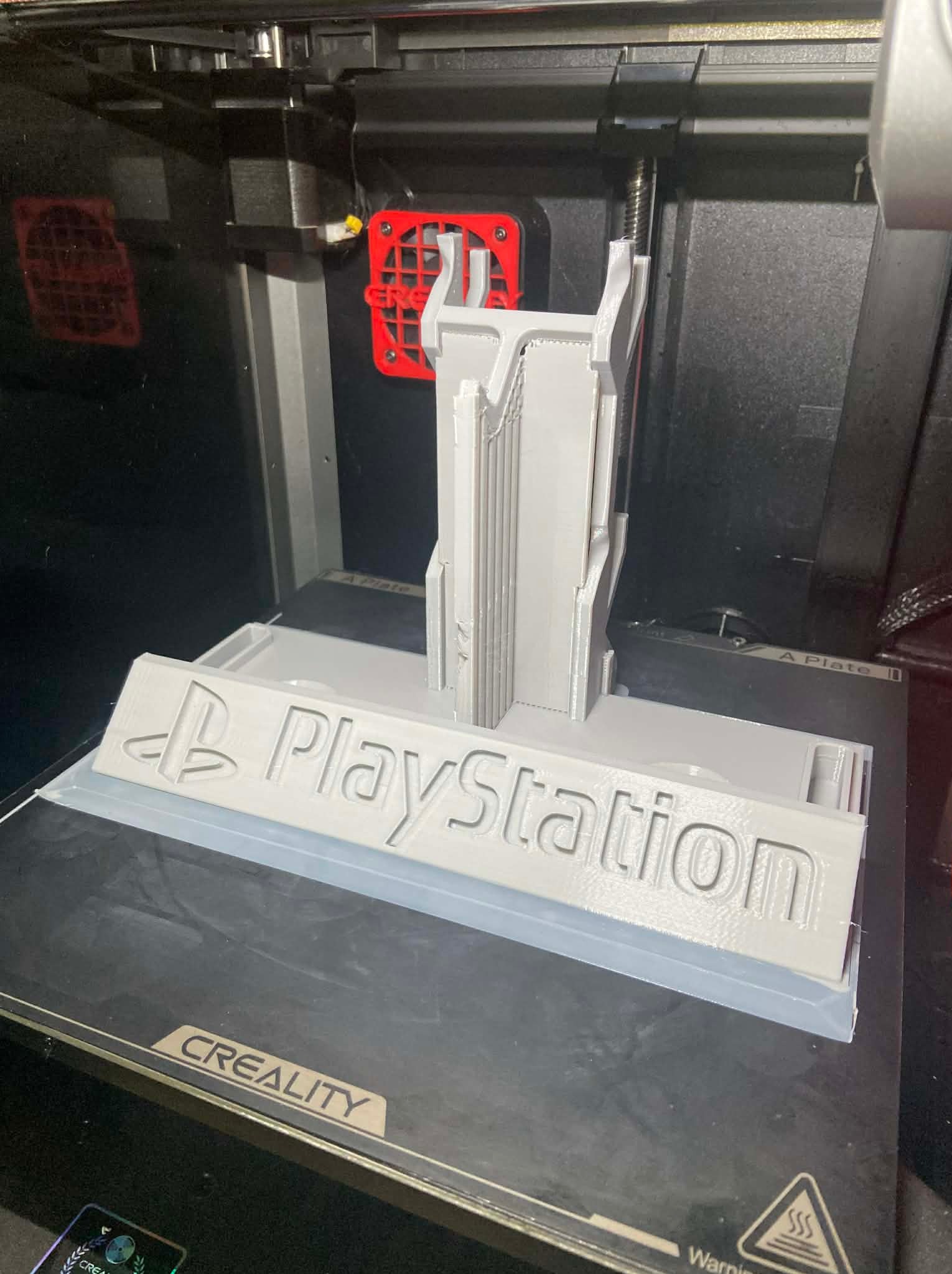 Szary model PlayStation z elementami architektonicznymi, powstający w drukarce 3D Creality. Widoczny wentylator chłodzący i platforma robocza drukarki.