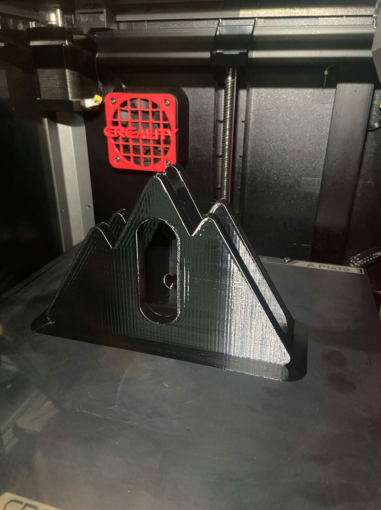 Czarny, trójwymiarowy model gór wydrukowany na drukarce 3D, widoczny wentylator drukarki. Detaliczne warstwy druku, ciemne tło wnętrza drukarki.