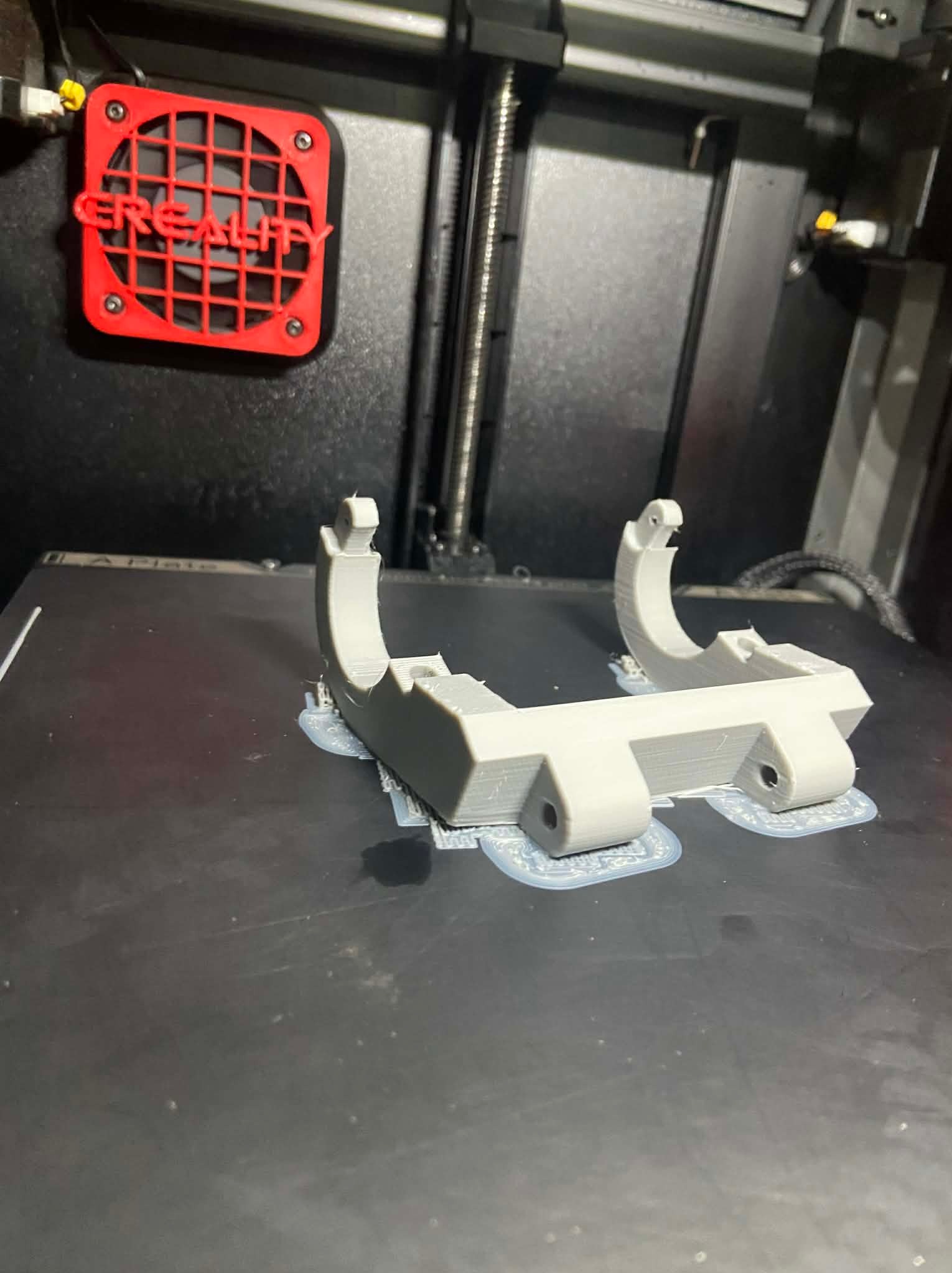 Szary, plastikowy element wydrukowany na drukarce 3D Creality, widoczny na czarnym stole drukarki. Detal z podporami, gotowy do odseparowania. Czerwony wentylator drukarki w tle.