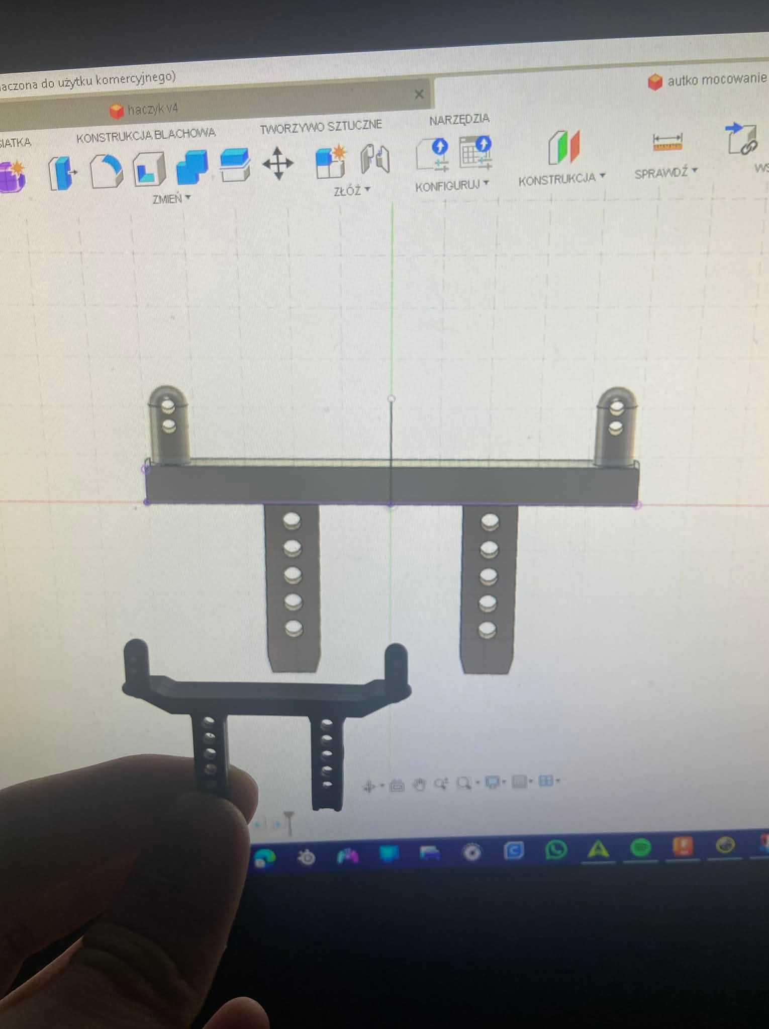 Model 3D haczyka w programie CAD, trzymany w dłoni, na tle ekranu z interfejsem projektowania. Widoczne narzędzia konstrukcyjne i nazwa pliku 'haczyk v4'.