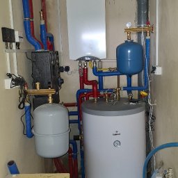 Piotr KAPŁON - Kotłownia z zamontowanym piecem gazowym, zbiornikiem na wodę i osprzętem. Widoczne rury w kolorach niebieskim i czerwonym. Ściany w surowym stanie.