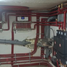 Piotr KAPŁON - Złożona instalacja grzewcza z czerwonymi rurami, rozdzielaczami i białym piecem Termet w piwnicy. Widoczne liczne przyłącza i elementy armatury.