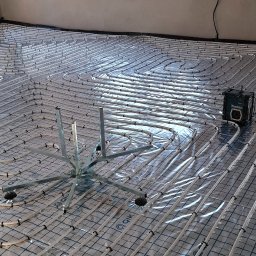 Piotr KAPŁON - Białe rury ogrzewania podłogowego ułożone na folii z niebieską siatką w surowym wnętrzu. Widoczne narzędzia montażowe i okno z widokiem na budynek.