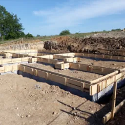 Szczegółowy widok z góry na szalunek fundamentów zbrojonych stalą, przygotowany do zalania betonem, na tle wykopu i błękitnego nieba.