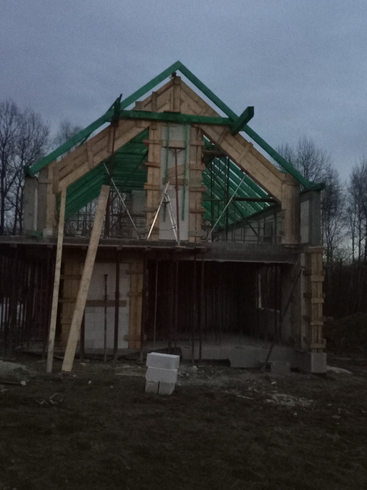 Surowy stan domu w trakcie budowy: widoczne szalunki, drewniana konstrukcja dachu z zielonymi elementami, bloczki betonowe na tle ponurego nieba i drzew.