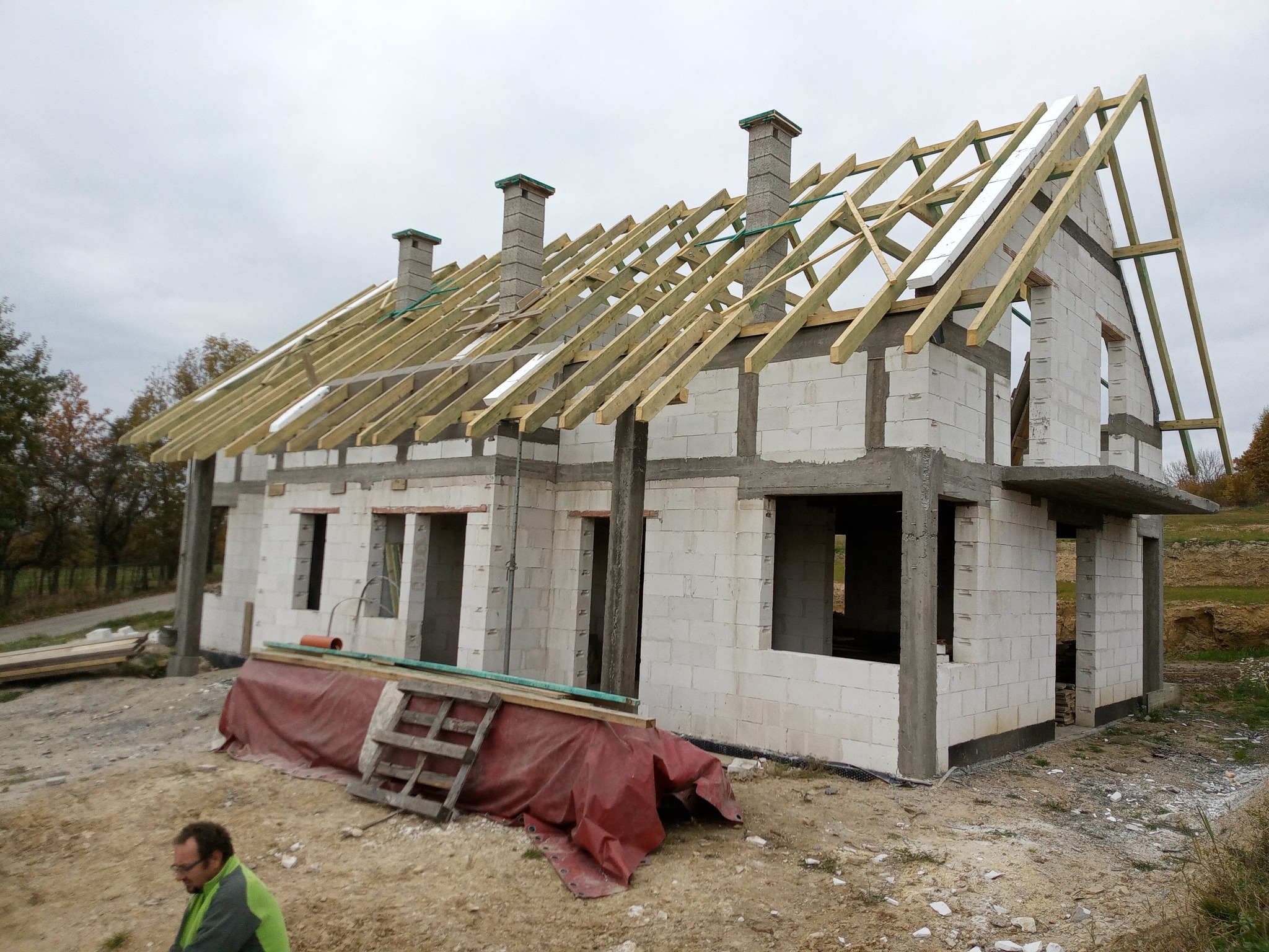 Wznoszenie domu z betonu komórkowego: surowe mury, więźba dachowa i trzy kominy w stanie surowym otwartym, widok z zewnątrz, częściowo zakryte materiały budowlane.