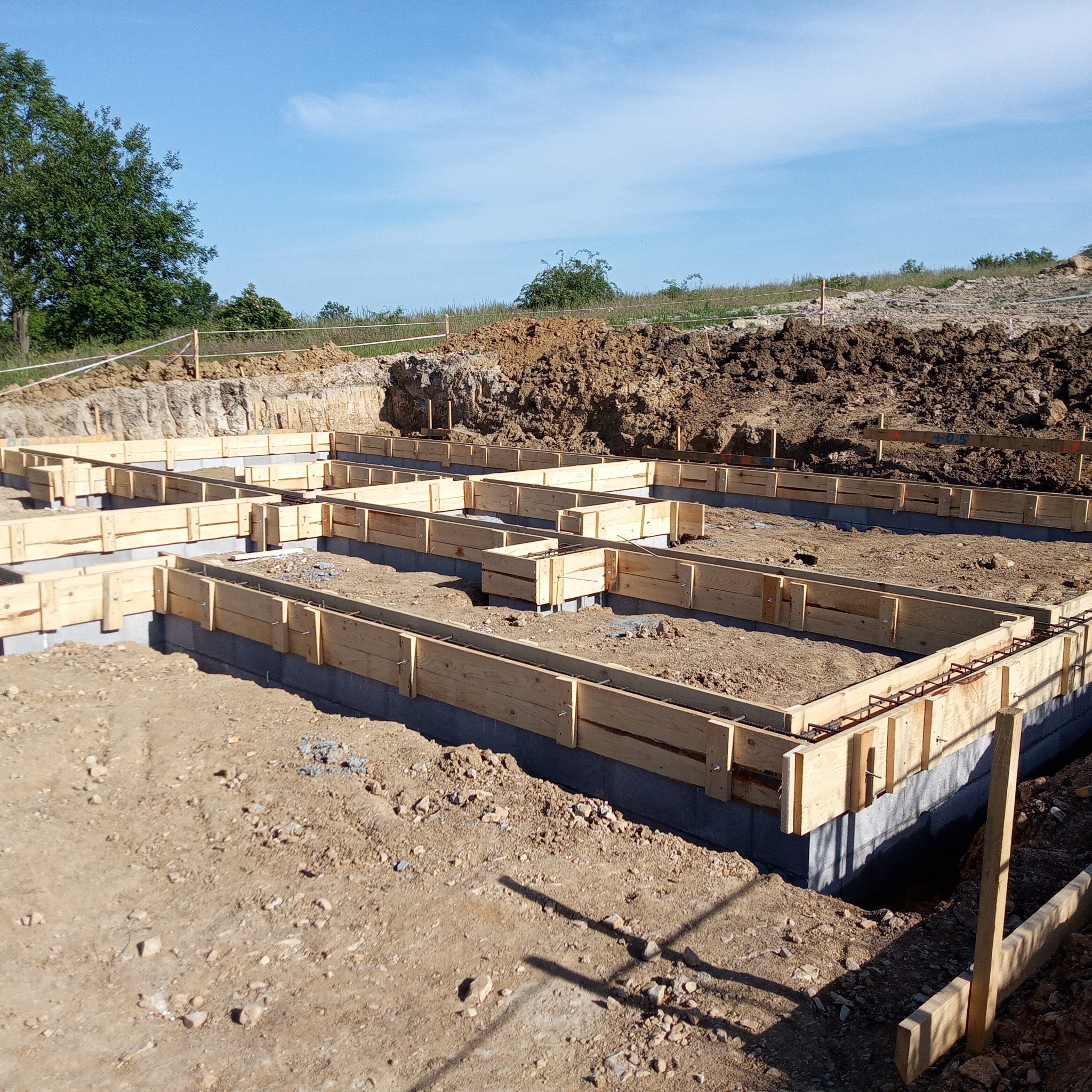 Szczegółowy widok z góry na szalunek fundamentów zbrojonych stalą, przygotowany do zalania betonem, na tle wykopu i błękitnego nieba.