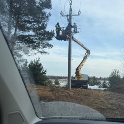ANISZEWSKI SP. Z O.O. - Prace na słupie energetycznym z podnośnika koszowego. Dwóch monterów w kaskach i kamizelkach. Fragment widoku z wnętrza pojazdu.