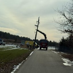 ANISZEWSKI SP. Z O.O. - Ekipa w trakcie wymiany słupa energetycznego przy drodze, widoczny dźwig i samochody serwisowe, zimowy krajobraz z resztkami śniegu.