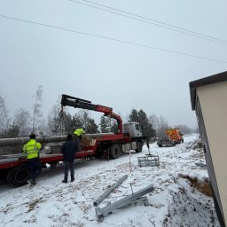 ANISZEWSKI SP. Z O.O. - Montaż słupów energetycznych za pomocą dźwigu na naczepie ciężarówki. Pracownicy w odblaskowych kamizelkach. Zimowy krajobraz.