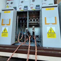 ANISZEWSKI SP. Z O.O. - Otwarta szafa elektryczna z widocznymi kablami i oznaczeniami ostrzegawczymi. Widoczne przewody uziemiające w oplocie miedzianym. Tabliczki informacyjne: Pole Nr 1 Liniowe.