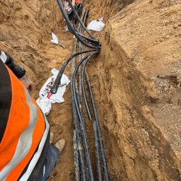 ANISZEWSKI SP. Z O.O. - Kable ułożone w wykopie ziemnym, częściowo zabezpieczone taśmą. Widoczne narzędzia i fragment osoby w odblaskowej kamizelce. Prace instalacyjne w toku.