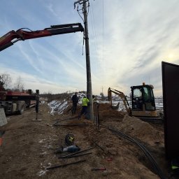 ANISZEWSKI SP. Z O.O. - Montaż słupa energetycznego z użyciem dźwigu i koparki. Widoczni pracownicy w kamizelkach odblaskowych, wykopany rów z kablami i narzędzia. Prace ziemne w pochmurny dzień.