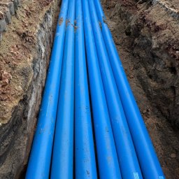 ANISZEWSKI SP. Z O.O. - Niebieskie rury ułożone w wykopie ziemnym, widoczne fragmenty nóg osoby stojącej nad wykopem. Instalacja podziemna w trakcie realizacji.