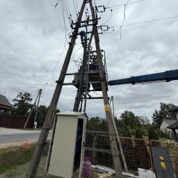 ANISZEWSKI SP. Z O.O. - Słup wysokiego napięcia z transformatorem i skrzynką rozdzielczą, widok z dołu na tle pochmurnego nieba. Widoczny podnośnik koszowy i butla gazowa.