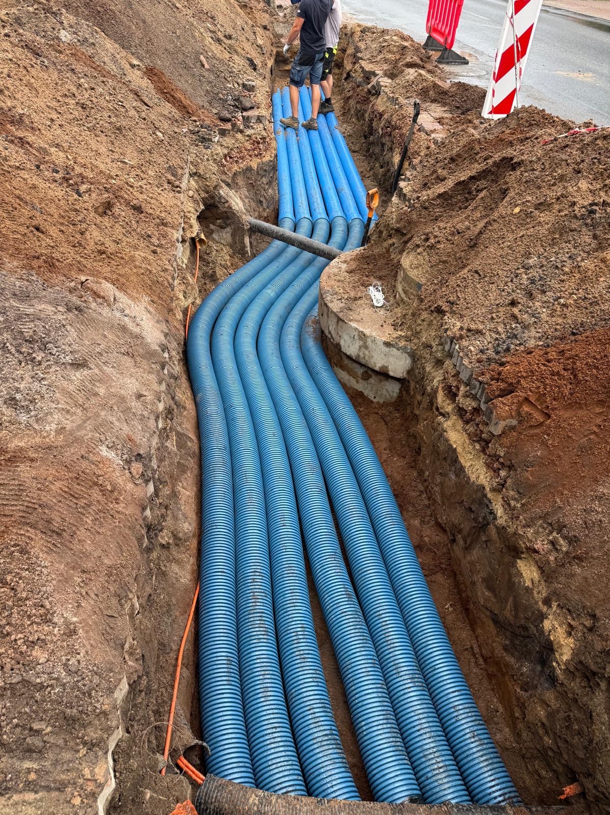 Wykop z ułożonymi niebieskimi rurami karbowanymi, prawdopodobnie pod kable. Widoczni pracownicy i fragment drogi. Prace ziemne w toku.