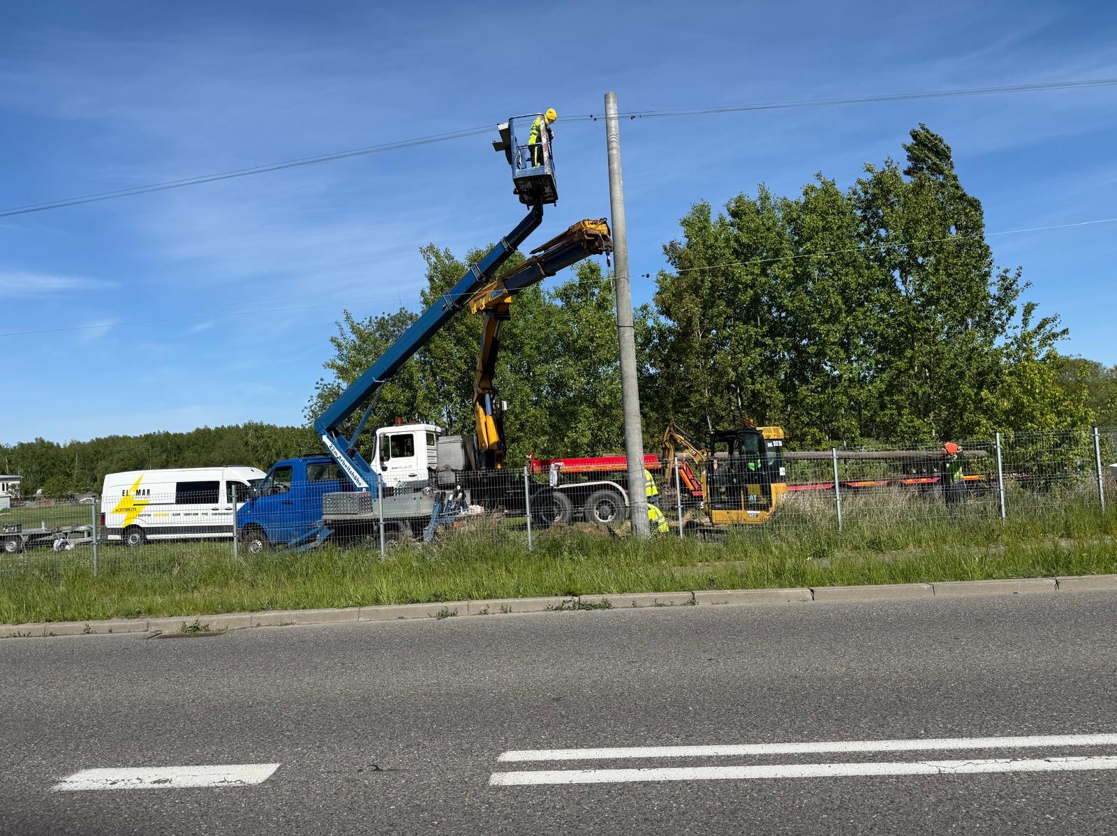 Prace przy słupie energetycznym z użyciem podnośnika koszowego. Widoczny pracownik w kasku i kamizelce. W tle zieleń i pojazdy. Dzień słoneczny.