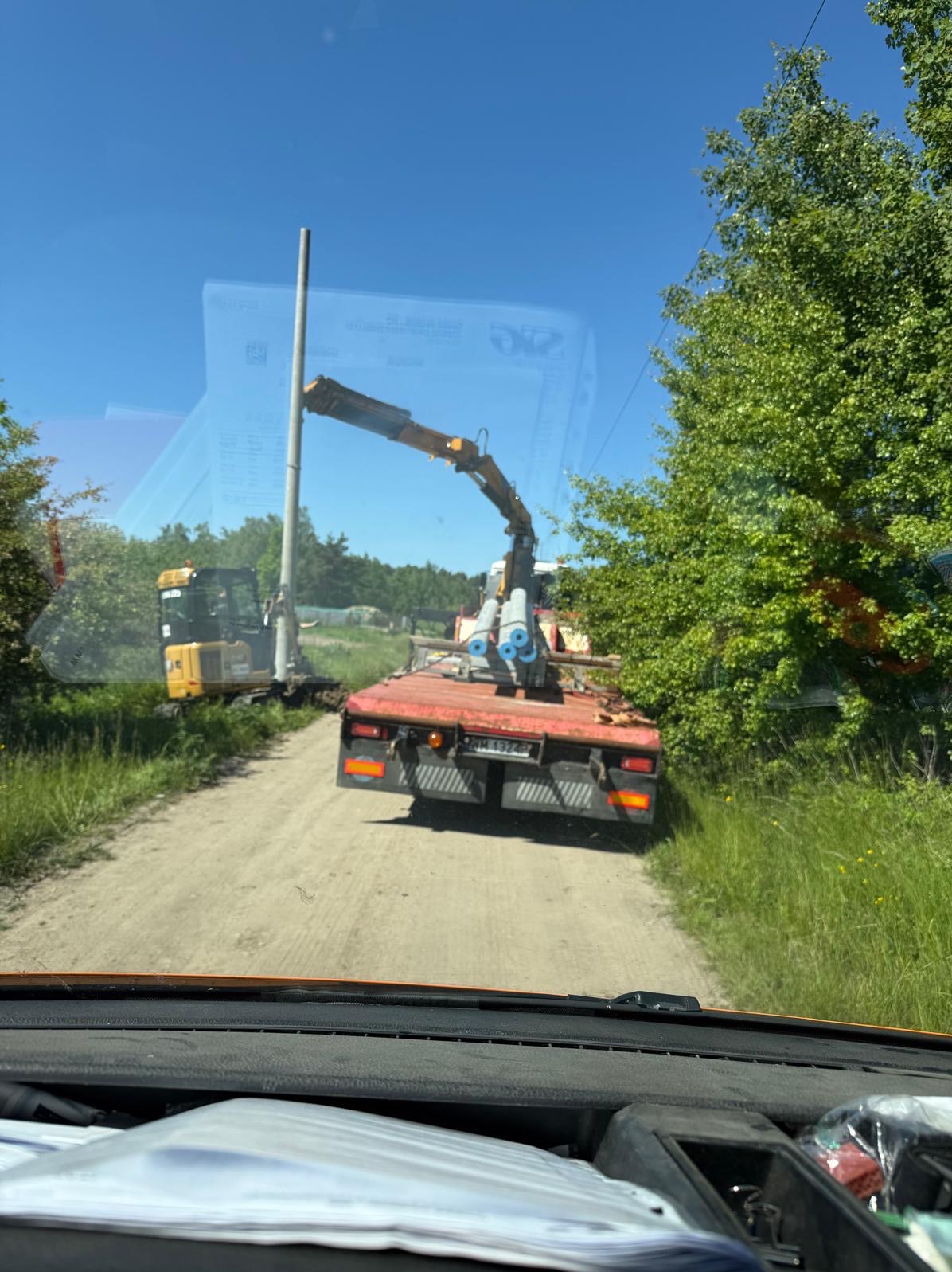 Transport słupów oświetleniowych na przyczepie z dźwigiem, obok koparka przy wykopanym dole. Widok z wnętrza pojazdu, słoneczny dzień.