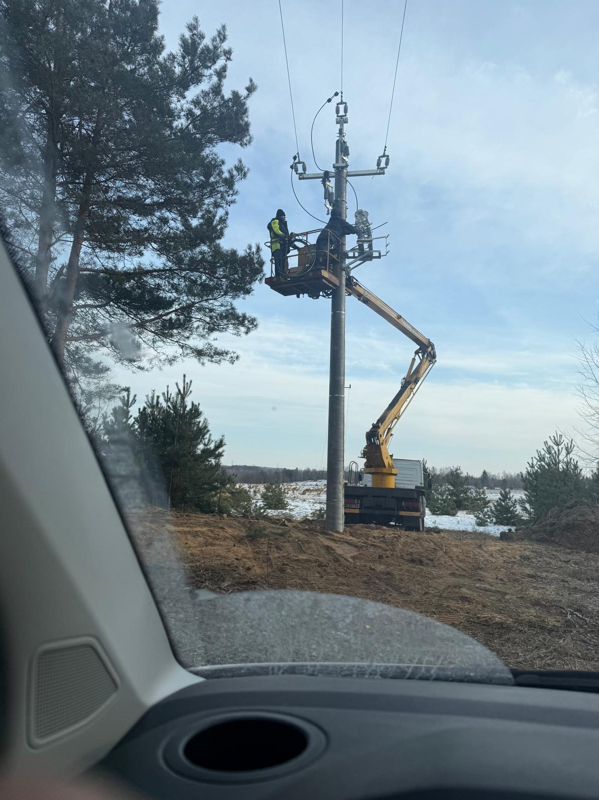 Prace na słupie energetycznym z podnośnika koszowego. Dwóch monterów w kaskach i kamizelkach. Fragment widoku z wnętrza pojazdu.