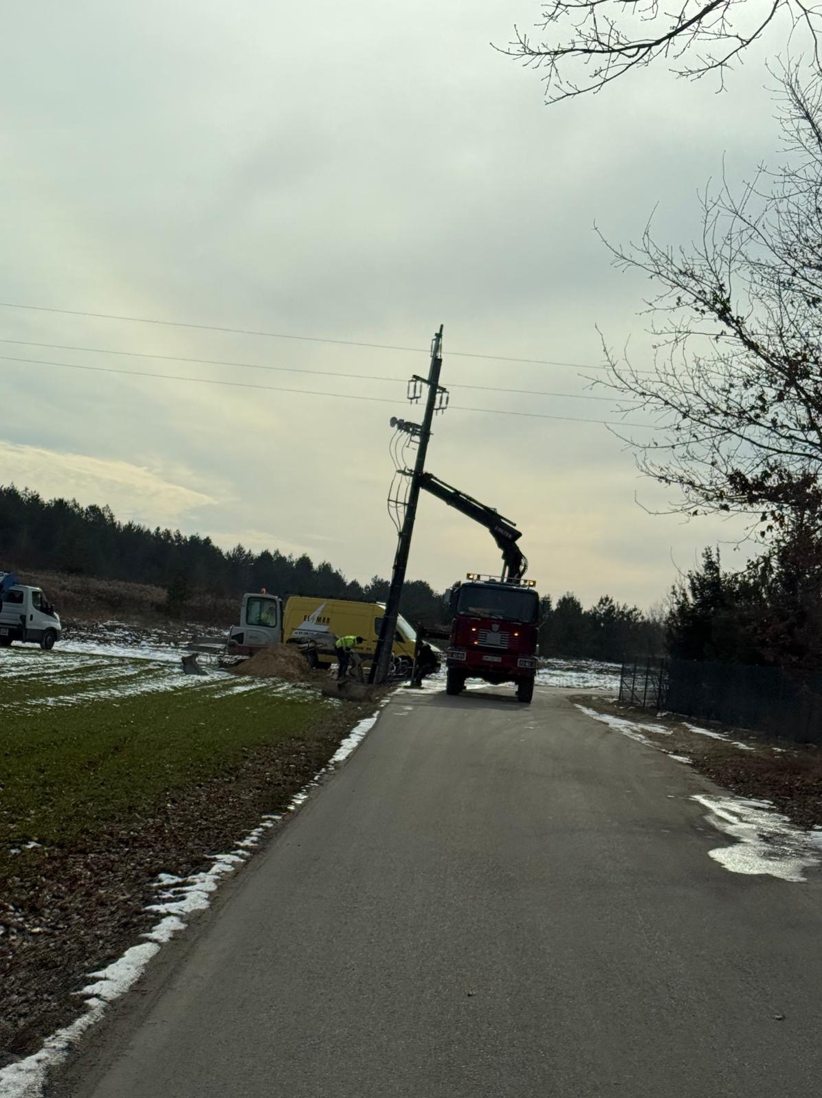 Ekipa w trakcie wymiany słupa energetycznego przy drodze, widoczny dźwig i samochody serwisowe, zimowy krajobraz z resztkami śniegu.