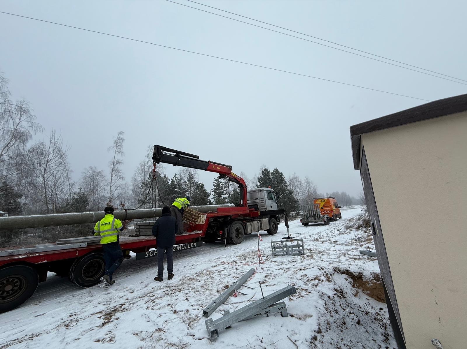 Montaż słupów energetycznych za pomocą dźwigu na naczepie ciężarówki. Pracownicy w odblaskowych kamizelkach. Zimowy krajobraz.