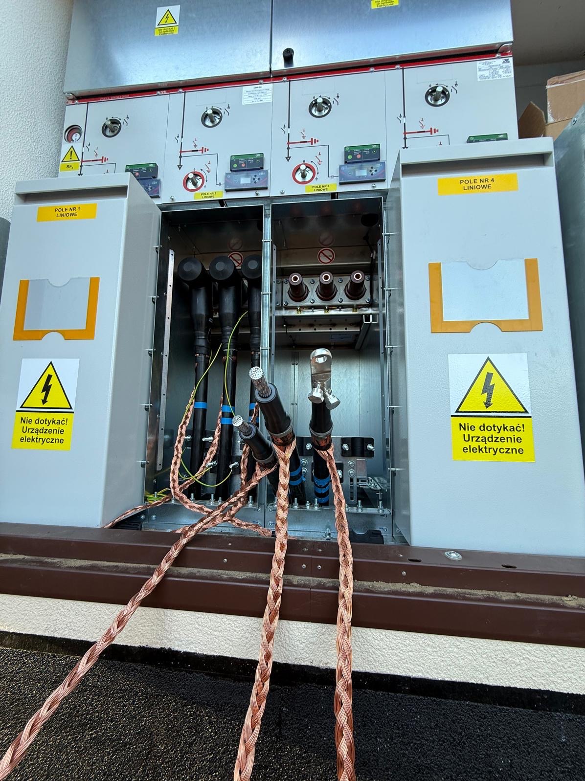 Otwarta szafa elektryczna z widocznymi kablami i oznaczeniami ostrzegawczymi. Widoczne przewody uziemiające w oplocie miedzianym. Tabliczki informacyjne: Pole Nr 1 Liniowe.