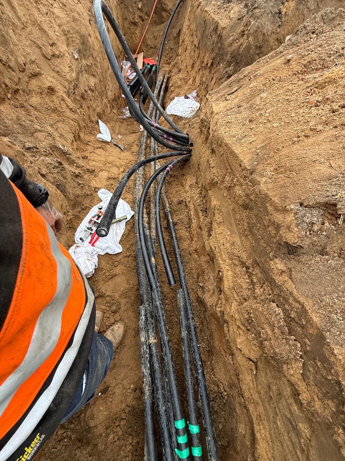 Kable ułożone w wykopie ziemnym, częściowo zabezpieczone taśmą. Widoczne narzędzia i fragment osoby w odblaskowej kamizelce. Prace instalacyjne w toku.