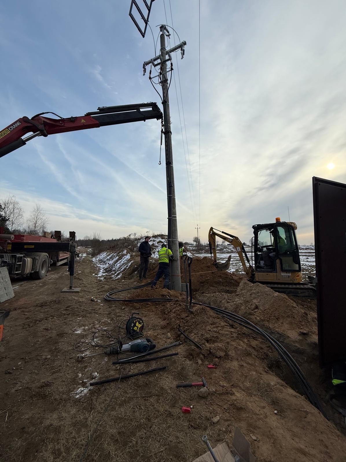 Montaż słupa energetycznego z użyciem dźwigu i koparki. Widoczni pracownicy w kamizelkach odblaskowych, wykopany rów z kablami i narzędzia. Prace ziemne w pochmurny dzień.
