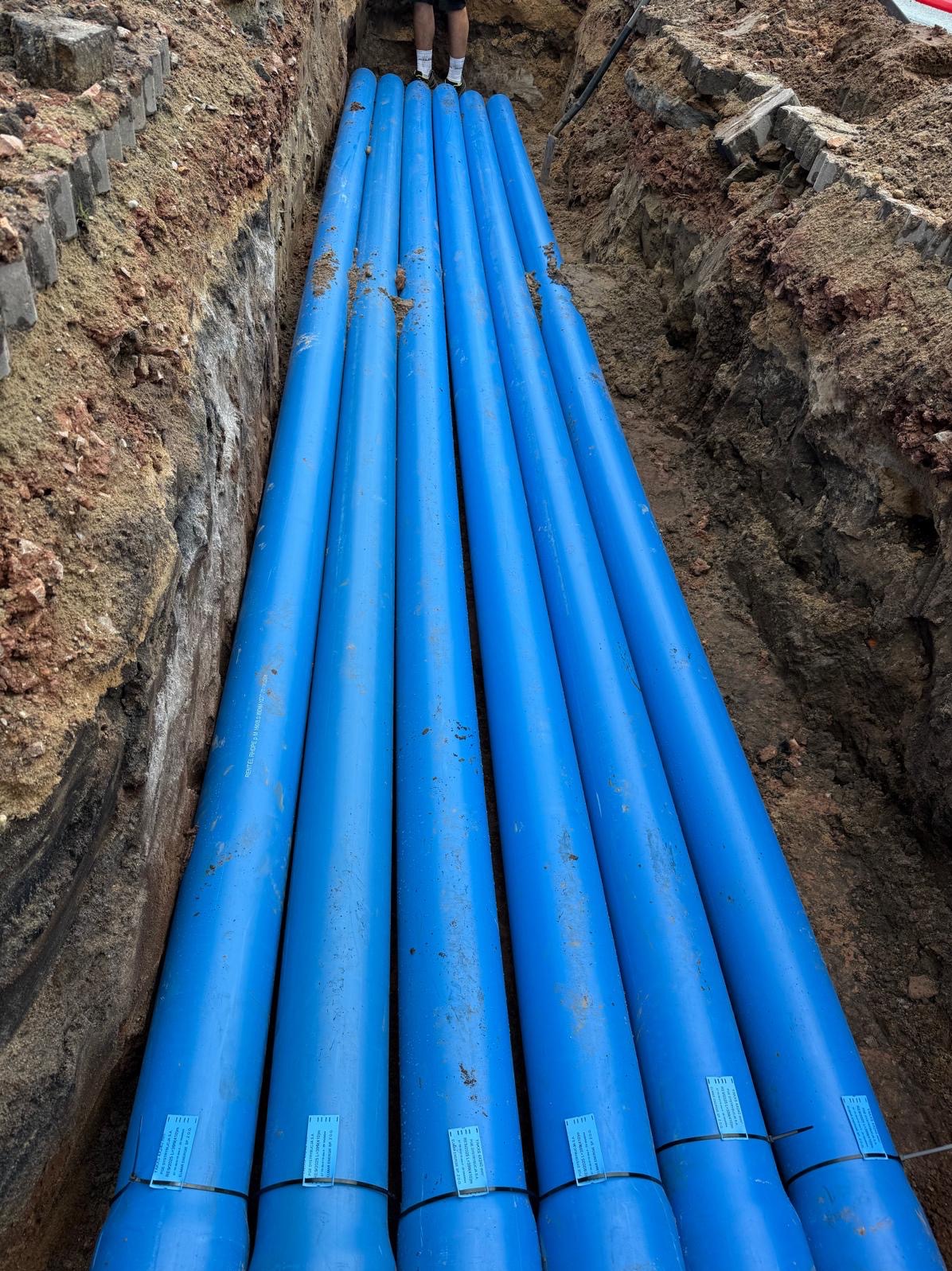 Niebieskie rury ułożone w wykopie ziemnym, widoczne fragmenty nóg osoby stojącej nad wykopem. Instalacja podziemna w trakcie realizacji.