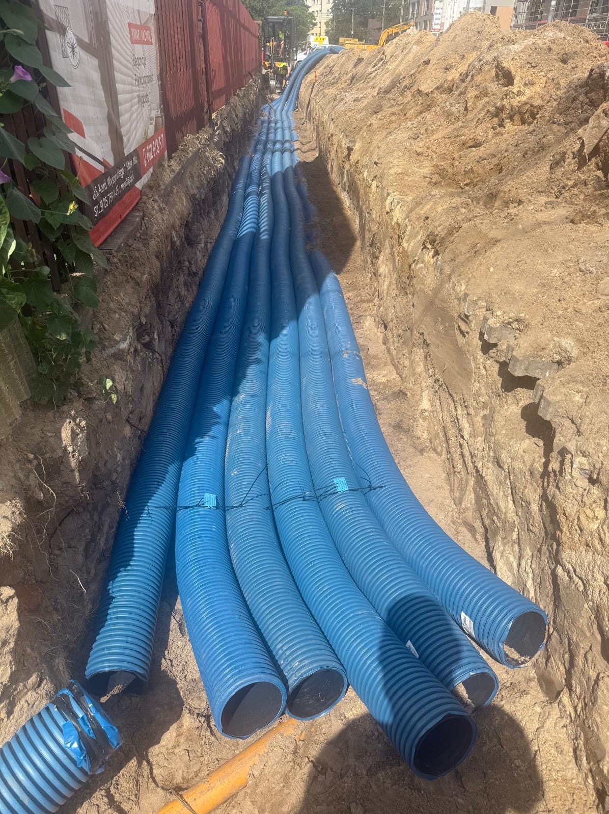 Wykop z ułożonymi niebieskimi rurami kablowymi, zabezpieczonymi taśmą. Widoczne ściany wykopu z ziemi i fragment budynku w tle. Prace ziemne w toku.