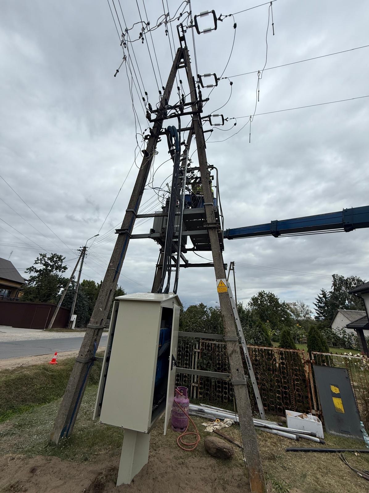 Słup wysokiego napięcia z transformatorem i skrzynką rozdzielczą, widok z dołu na tle pochmurnego nieba. Widoczny podnośnik koszowy i butla gazowa.