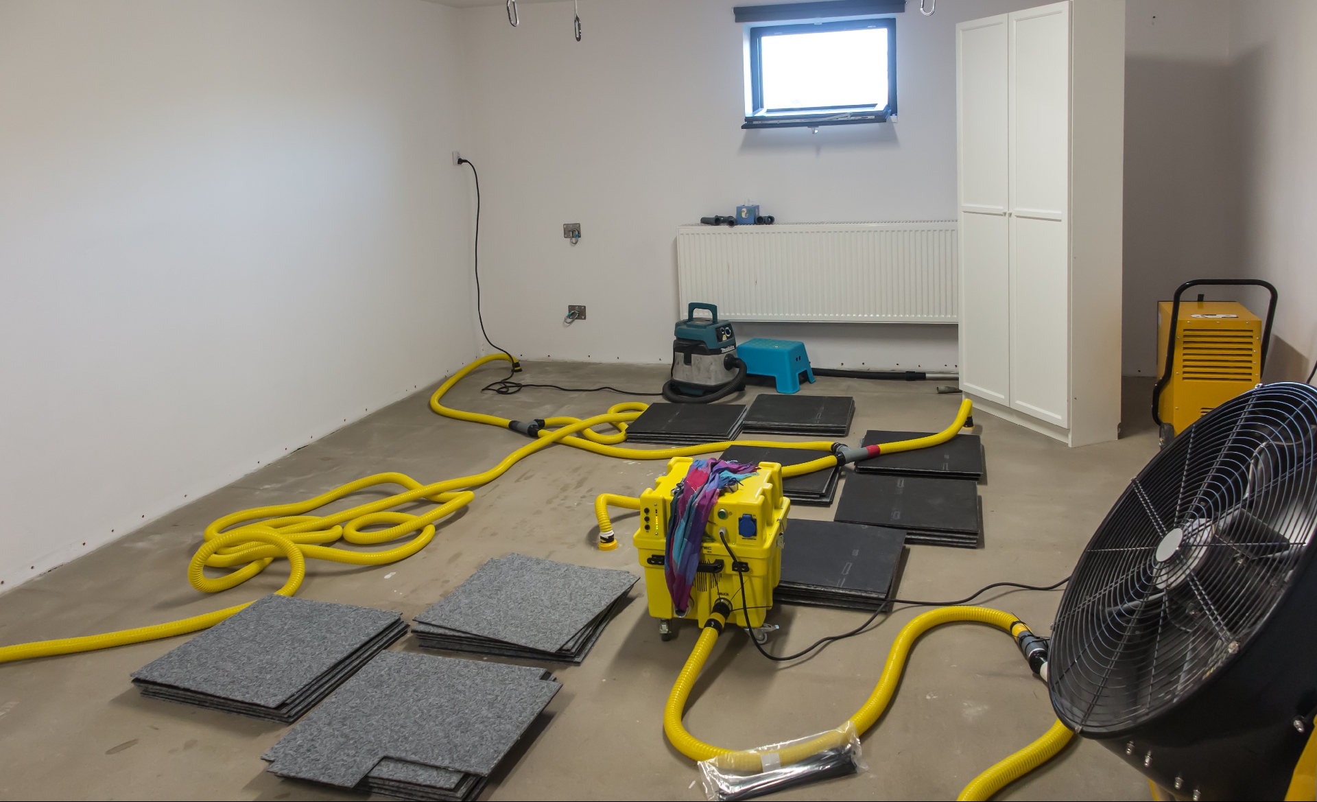 Żółte węże i czarne maty na podłodze w pomieszczeniu z oknem i grzejnikiem. Sprzęt do osuszania w trakcie pracy. Dodatkowo widoczny wentylator i odkurzacz.