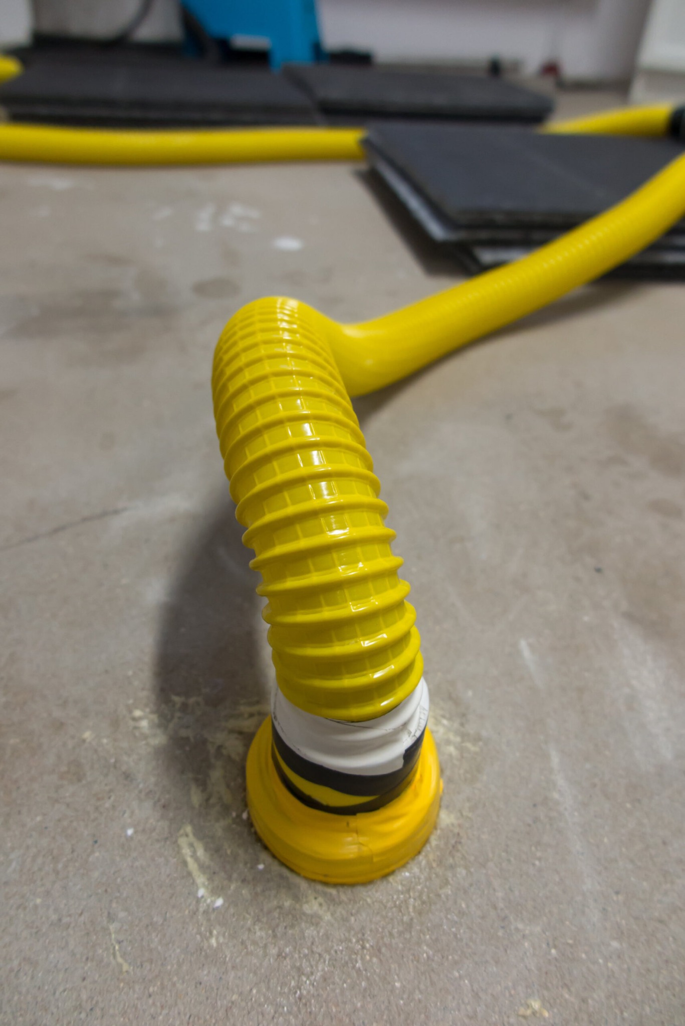 Żółty, karbowany wąż wentylacyjny przymocowany do podłoża, prawdopodobnie w trakcie procesu usuwania wilgoci. Widoczne płyty w tle.